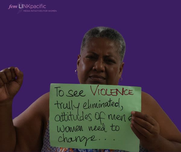 A woman is raising her fist while holding up a sign that reads, "To see violence truly eliminated, attitudes of men and women need to change..."
