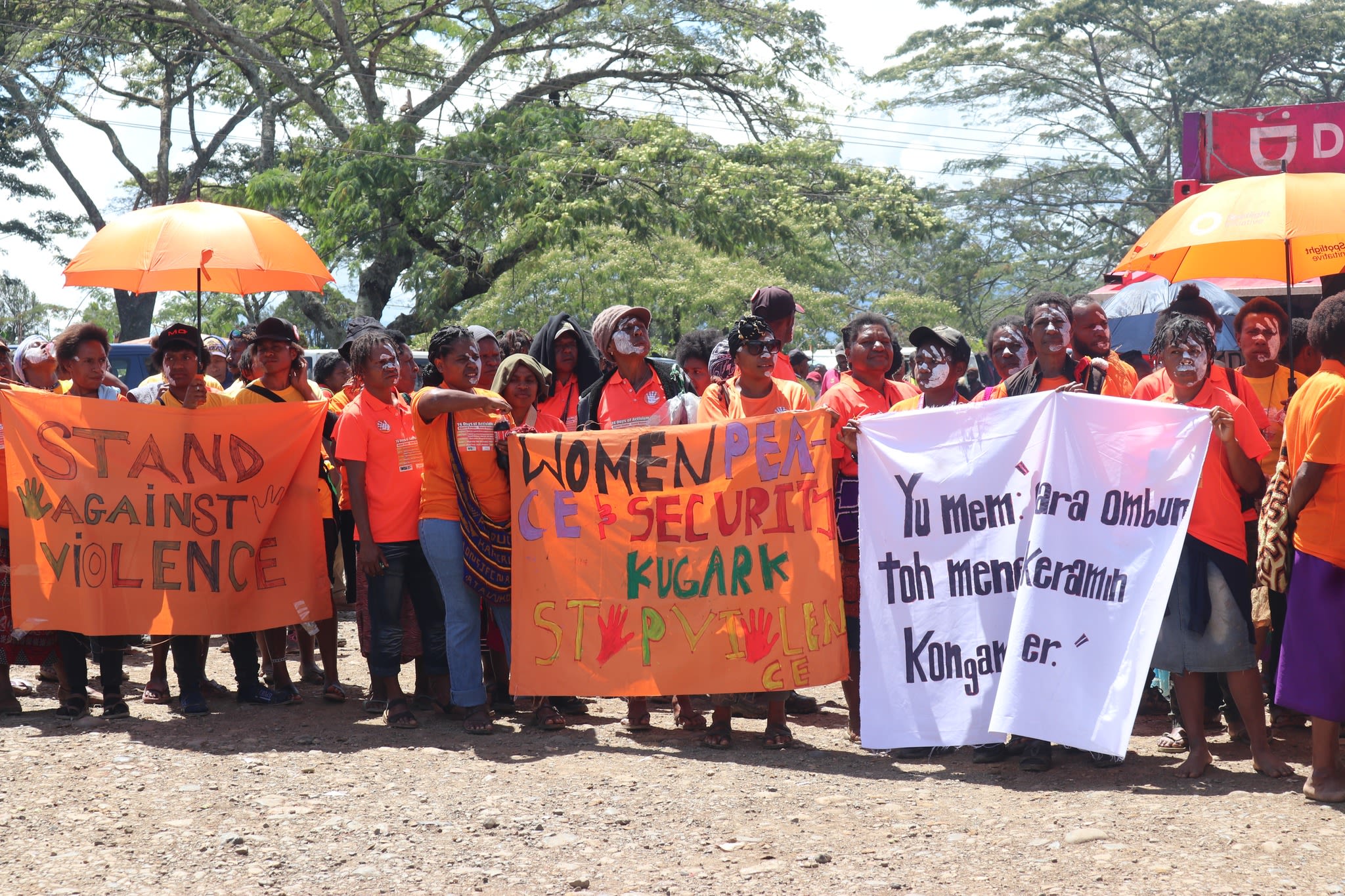 A crowd wearing orange t-shirts behind a series of handmade banners with messages about ending violence against women and girls.