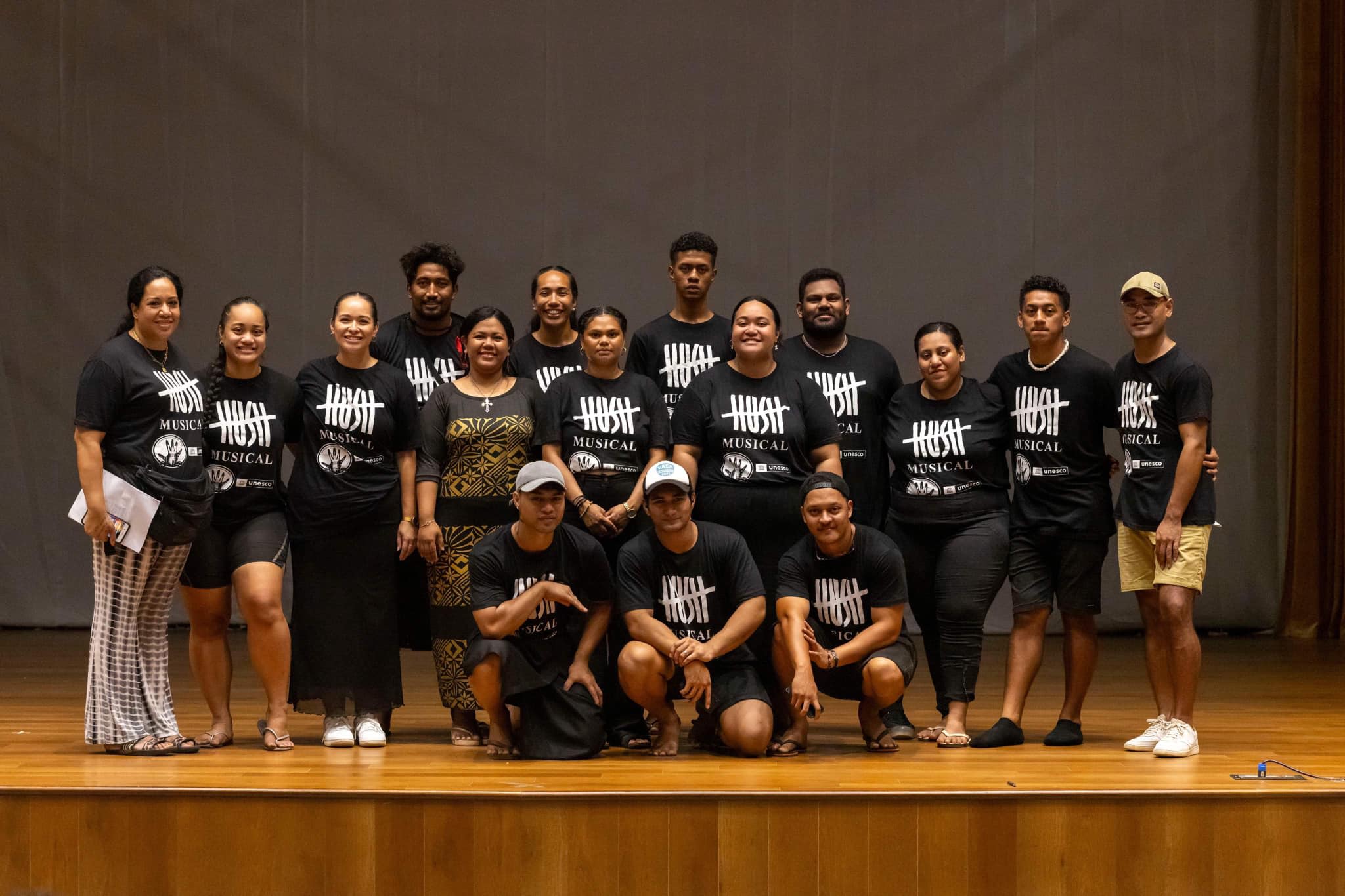 The cast and crew of Hush, all wearing black t-shirts with the musical's title on them', are posing together on stage.