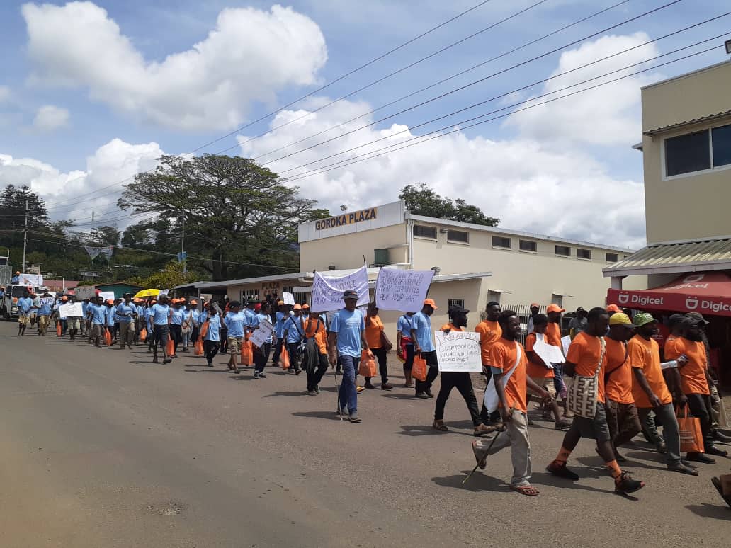 A crowd is marching through the streets of Goroka Town. They are holding banners and wearing blue and orange shirts.