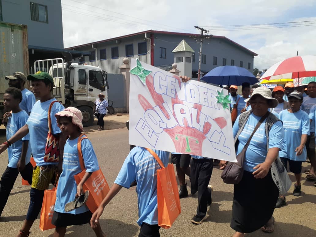 A group of people wearing blue t-shirts and carrying orange tote bags marching through the streets of Goroka Town. One of them is holding up a banner that reads, "Gender Equality."