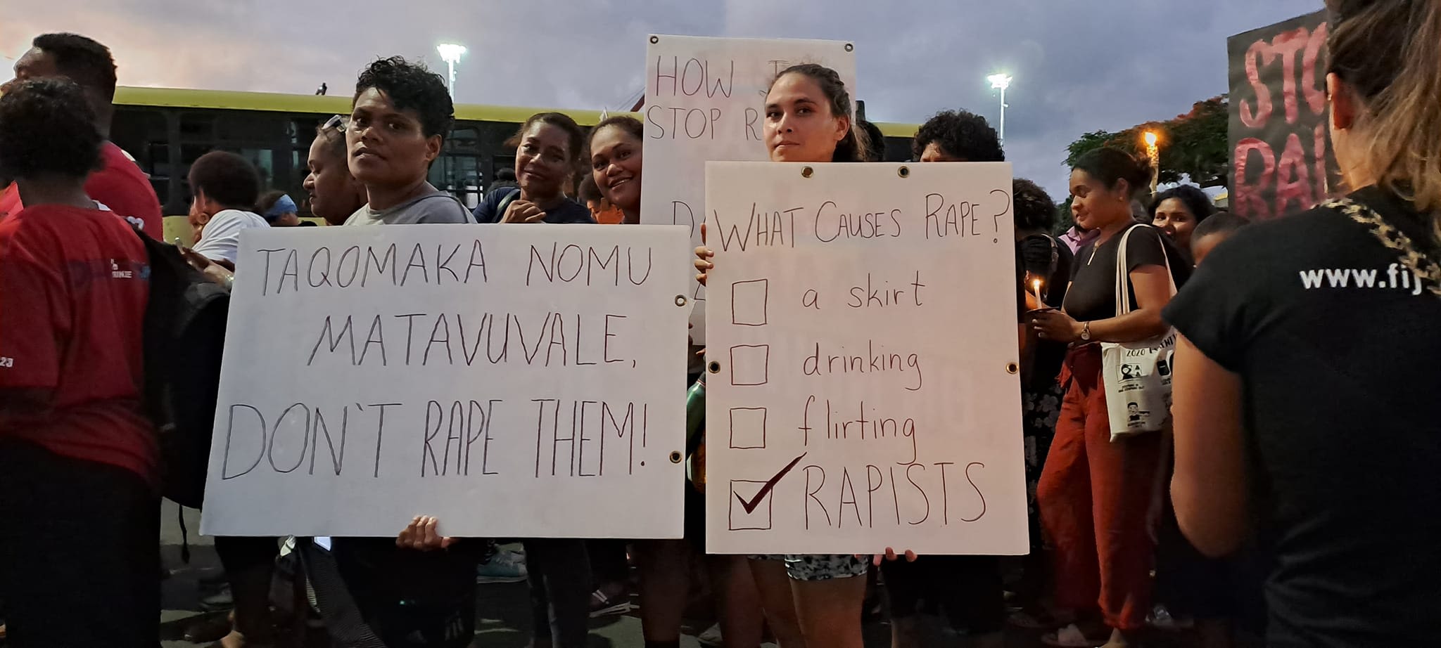 People looking to the camera as they hold up signs at the Reclaim the Night march. One poster reads, 'What causes rape?'. Below are empty checkboxes that read, 'a skirt, drinking, flirting', with the only ticked checkbox saying, 'rapists'.
