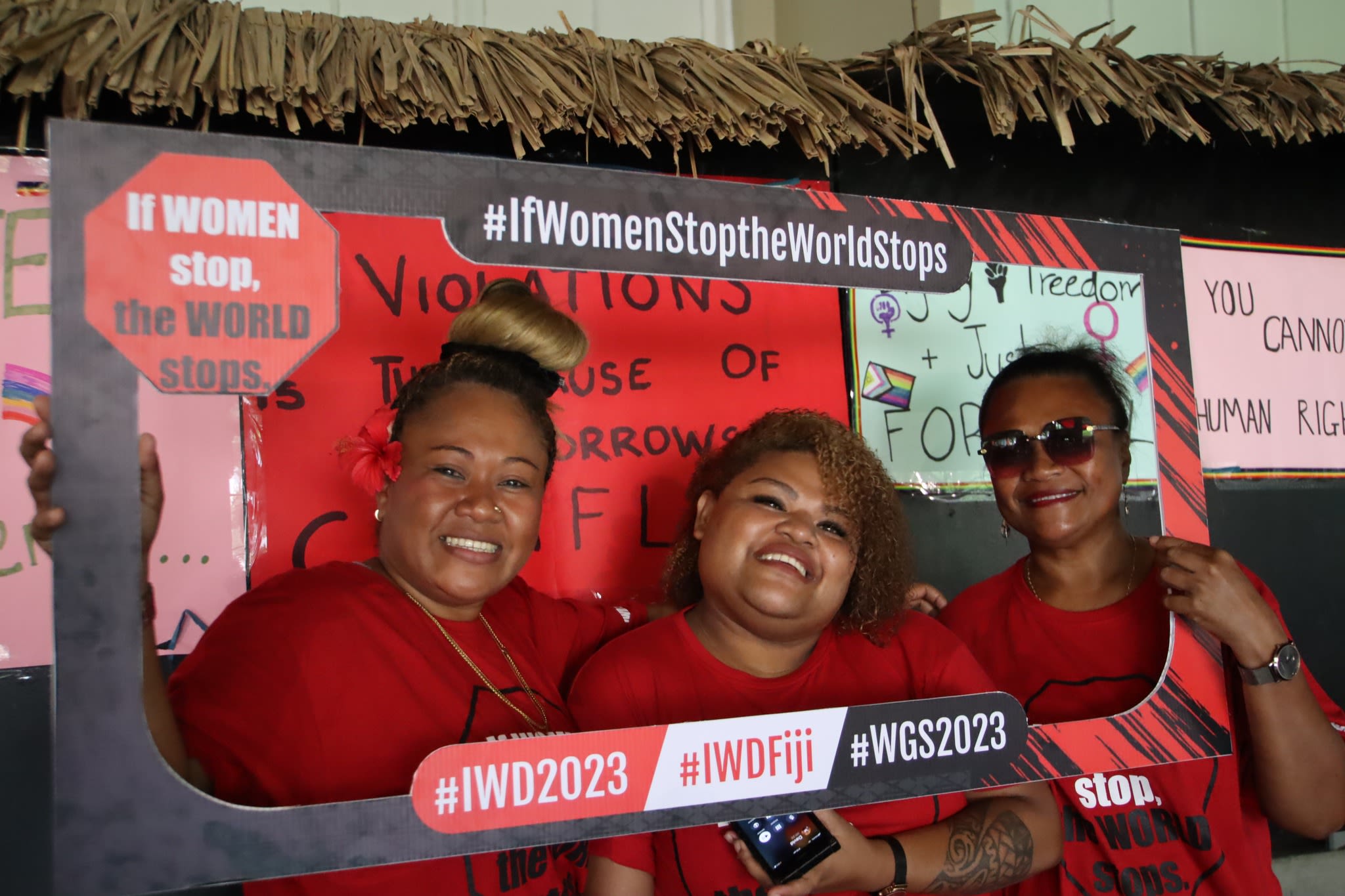 Three women dressed in red t-shirts smiling at the camera as they pose with a selfie frame that reads, 'If Women Stop, The World Stops - #IWD2023 #IWDFiji #WGS2023'. 