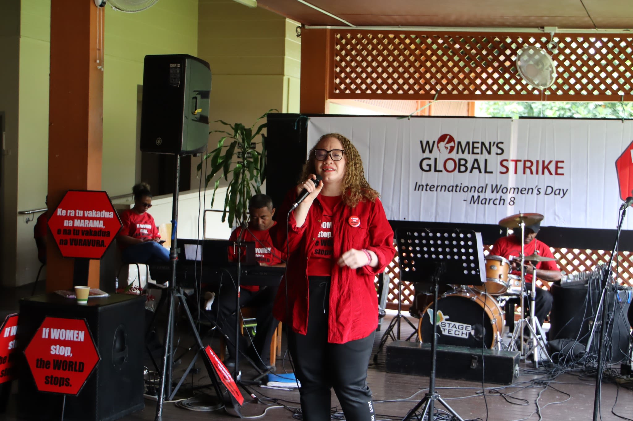 A woman dressed in a red t-shirt and shirt speaking on stage. Behind her is a banner that reads, 'Women's Global Strike: International Women's Day - March 8'.
