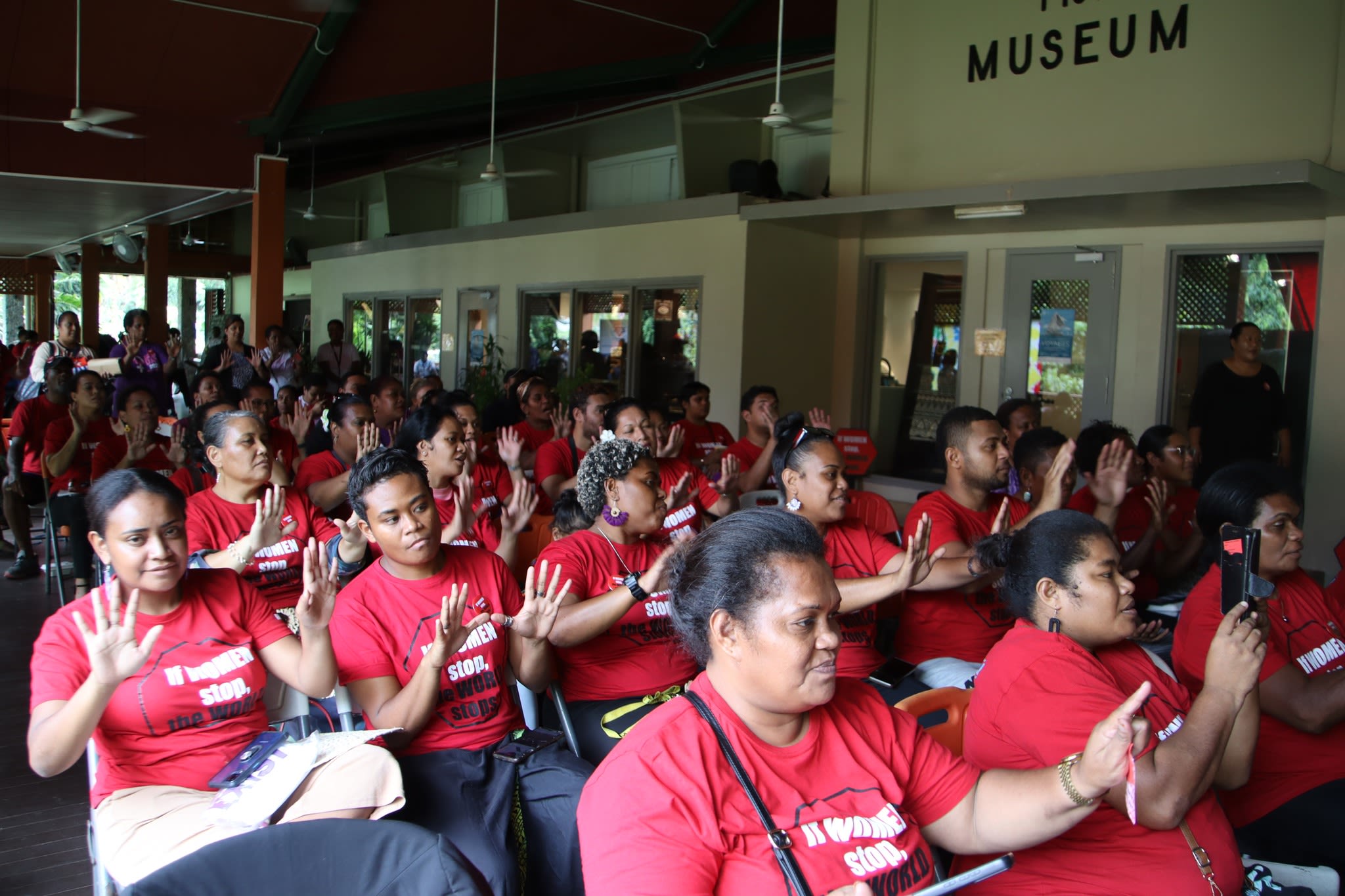 A seated crowd of people wearing matching 'If Women Stop, The World Stops' red t-shirts. They are holding up their hands and looking towards the stage.