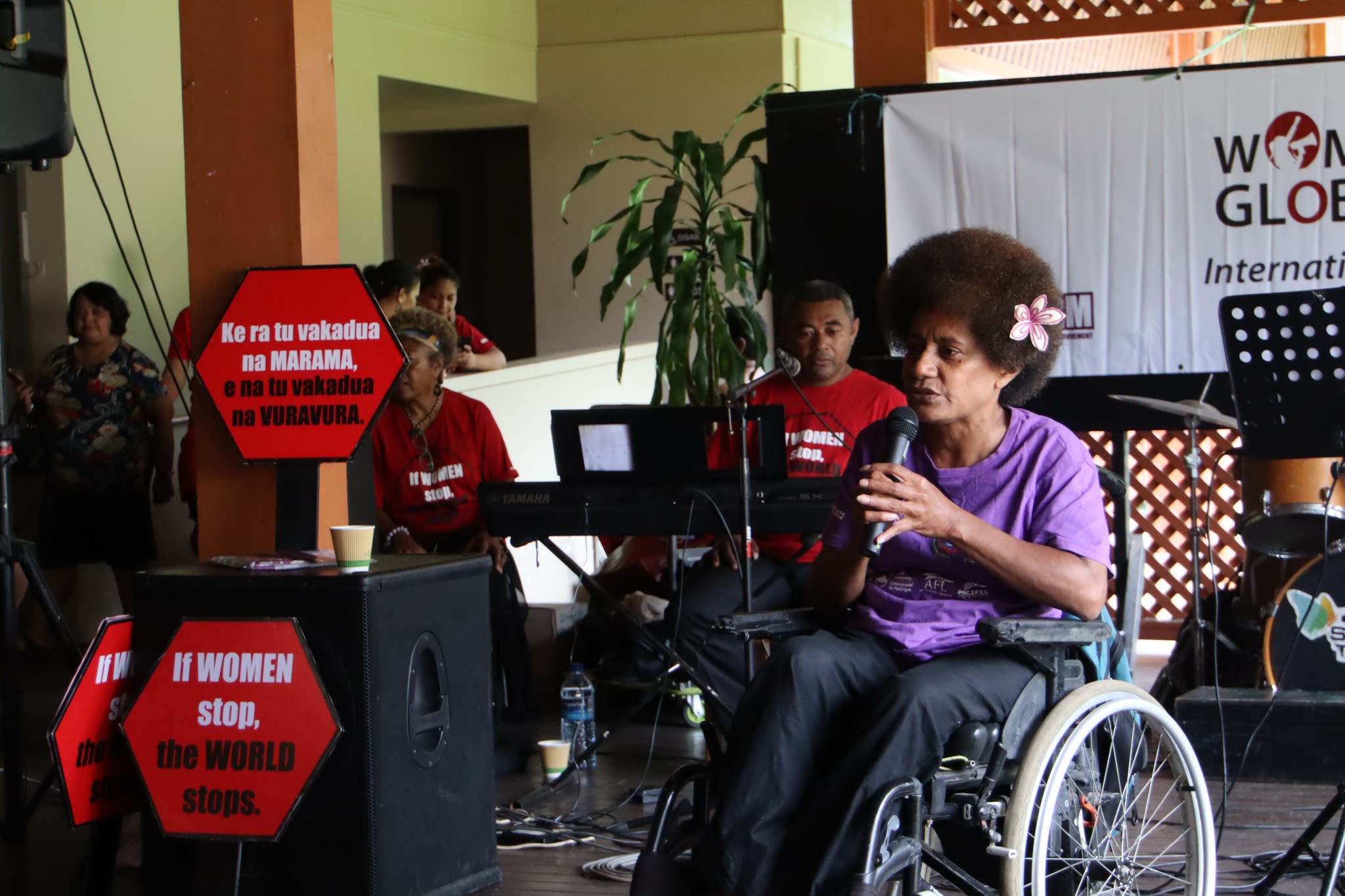 A woman in a wheelchair wearing a purple t-shirt and matching flower in her hair speaking on stage.
