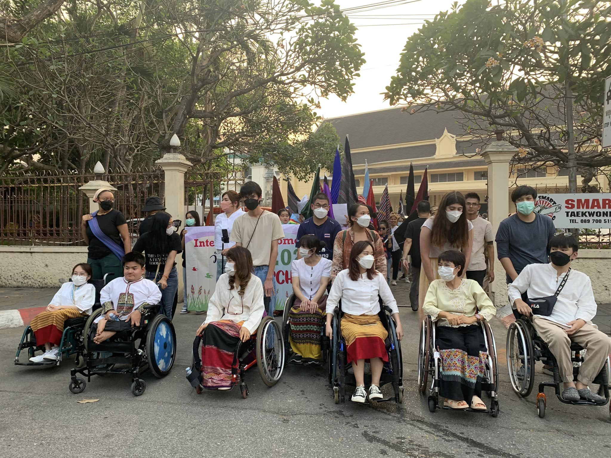 People participating in Women's League of Burma's International Women's Day march. Some are wheelchair users with people carrying banners and flags behind them.